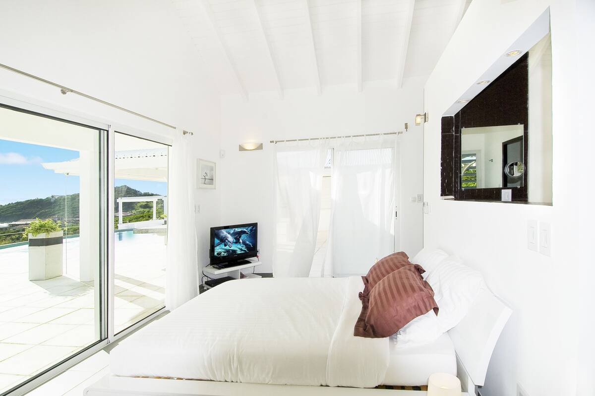 A modern bedroom with a large bed, a TV, and a balcony with a view of the landscape.