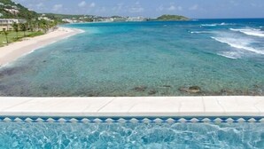 A clear view of a swimming pool with a tiled edge, overlooking a beach with clear turquoise water and a distant island. - Beach House Younes by Island Properties Online (Oyster Pond)