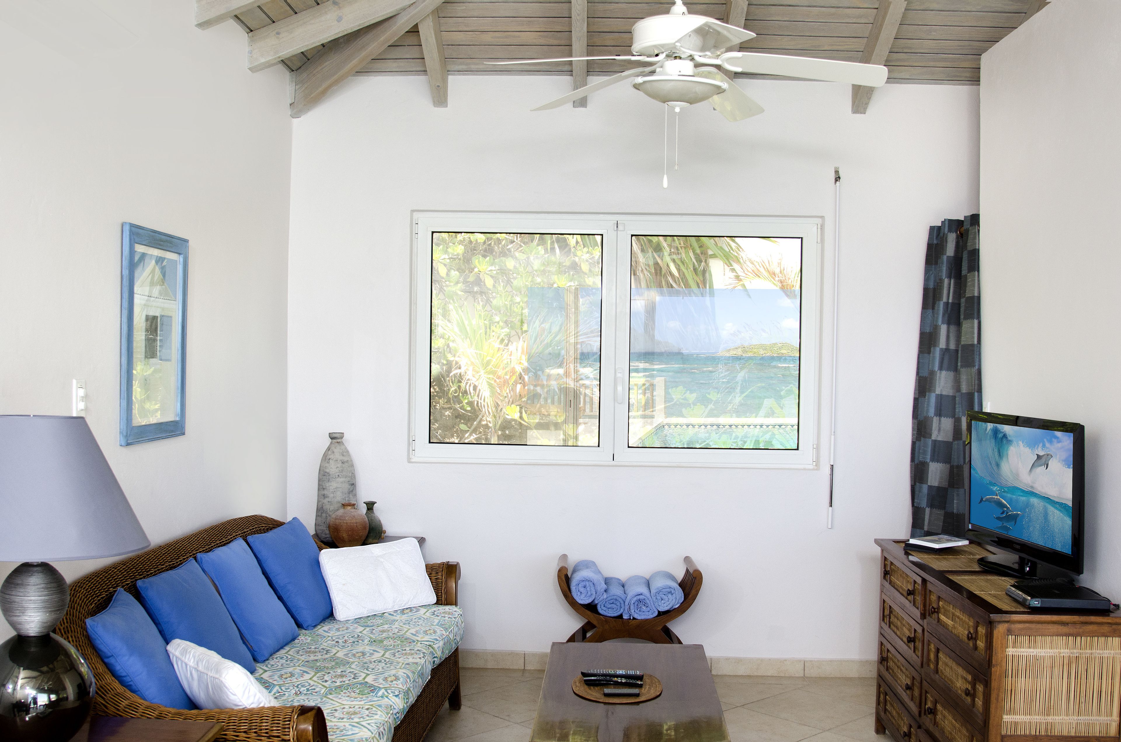 Una habitación con ventilador de techo, un sofá con cojines azules, un televisor y vistas al océano a través de amplias ventanas.