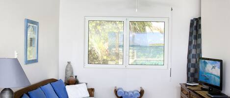 Una habitación con ventilador de techo, un sofá con cojines azules, un televisor y vistas al océano a través de amplias ventanas.