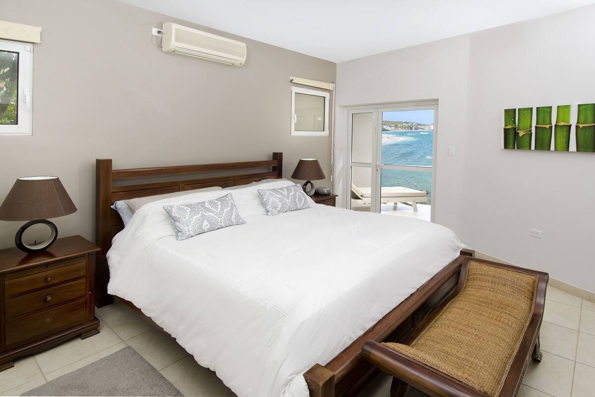 A bedroom with a bed, bedside tables, a lamp, a chair, and a view of the sea.