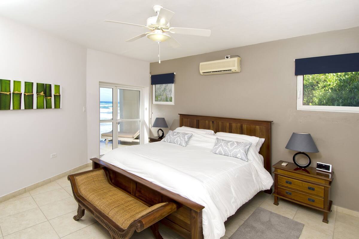 A bedroom with a bed, a wooden nightstand, a ceiling fan, a window with a view, and a door leading to a balcony.