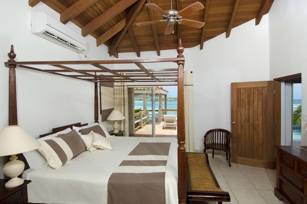 Un lit à baldaquin avec un ventilateur de plafond, une armoire en bois et une vue sur la plage, visible à travers une porte vitrée.