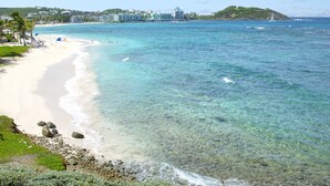 Une plage à l’eau turquoise et limpide, une rive sablonneuse et une vue lointaine sur un complexe hôtelier composé de plusieurs bâtiments.