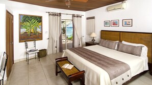Une chambre d’hôtel avec un lit, un ventilateur de plafond, une fenêtre avec des rideaux, un tableau et une vue sur l’océan.