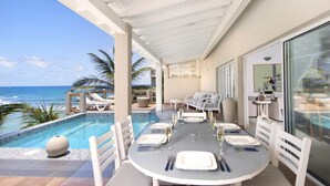 Un espace repas aménagé au bord de la piscine, avec vue sur l’océan, une table dressée pour le repas, et un coin salon avec un canapé et des fauteuils.