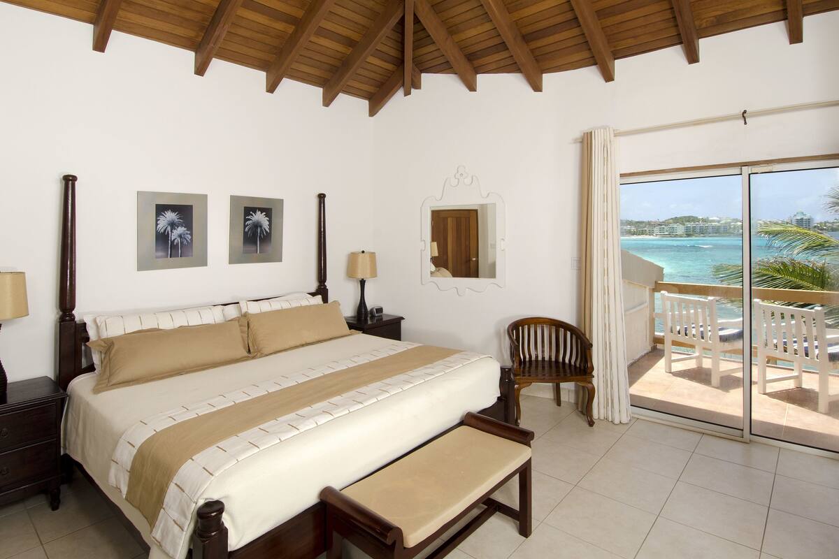 Une chambre avec un grand lit, un banc, une chaise, une table de chevet, une lampe et une vue sur l’océan par une porte-fenêtre coulissante.