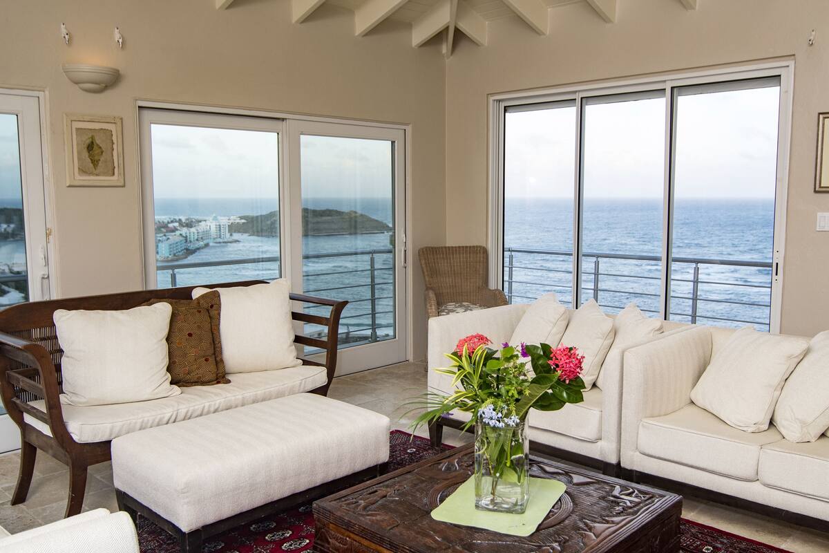 A living room with a sofa, a coffee table with a vase of flowers, and large windows offering a view of the ocean.