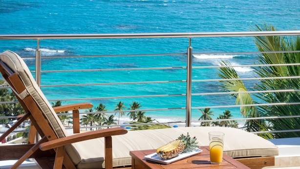 Uma varanda com vista para o oceano, uma cadeira de madeira, um travesseiro e uma mesa com uma abacaxi e um copo de suco de laranja.