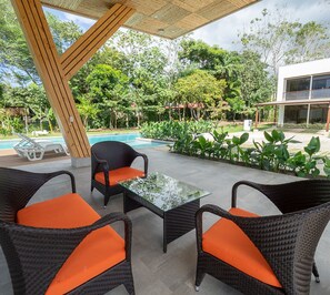 An outdoor seating area with wicker chairs and a glass table, overlooking a swimming pool and lush greenery.
