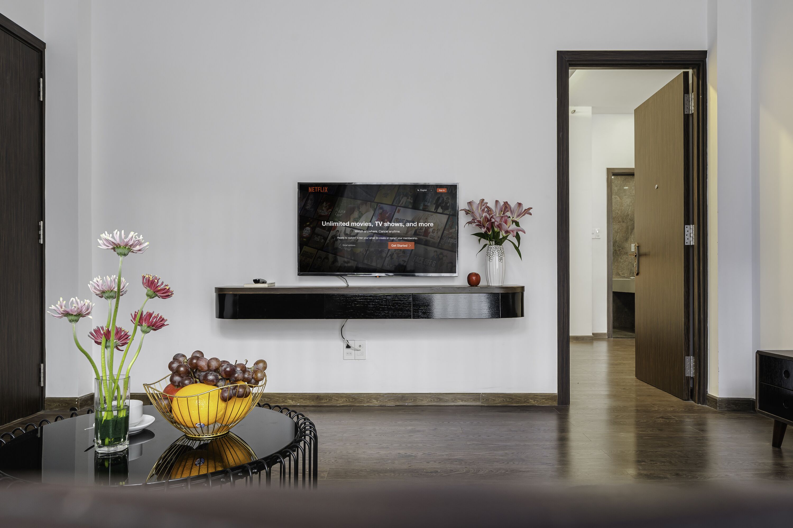 A modern living room with a flat-screen TV mounted on a wall, a dining table with a vase of flowers, and a hallway leading to another room.