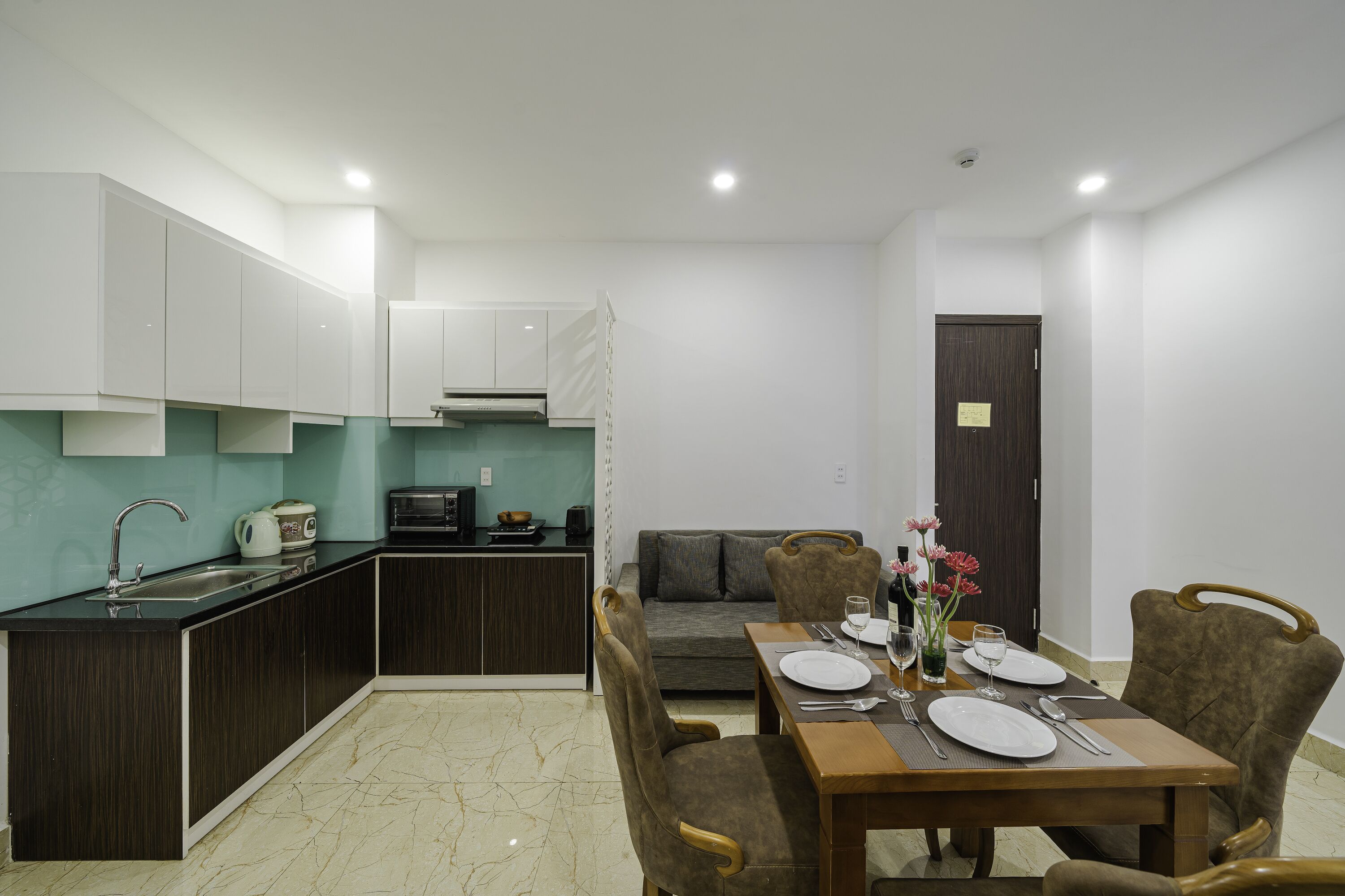 A modern kitchen with dark cabinetry, a dining area with a wooden table set for four, and a grey sofa in the background.