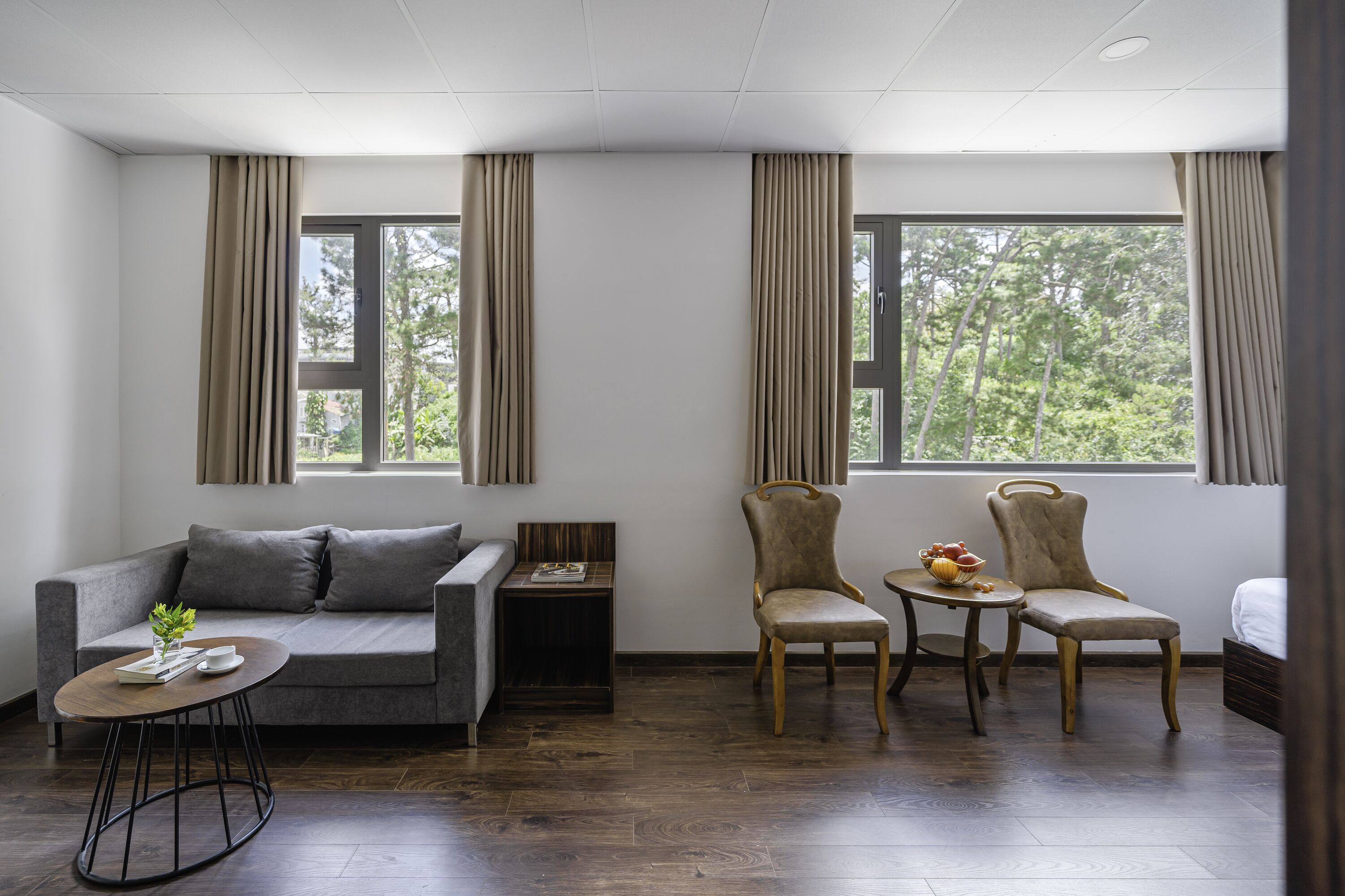Una habitación moderna con un sofá gris, dos sillones, una pequeña mesa redonda y una mesita de madera. Grandes ventanales con cortinas ofrecen vistas a árboles.