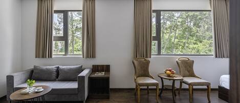 Una habitación moderna con un sofá gris, dos sillones, una pequeña mesa redonda y una mesita de madera. Grandes ventanales con cortinas ofrecen vistas a árboles.
