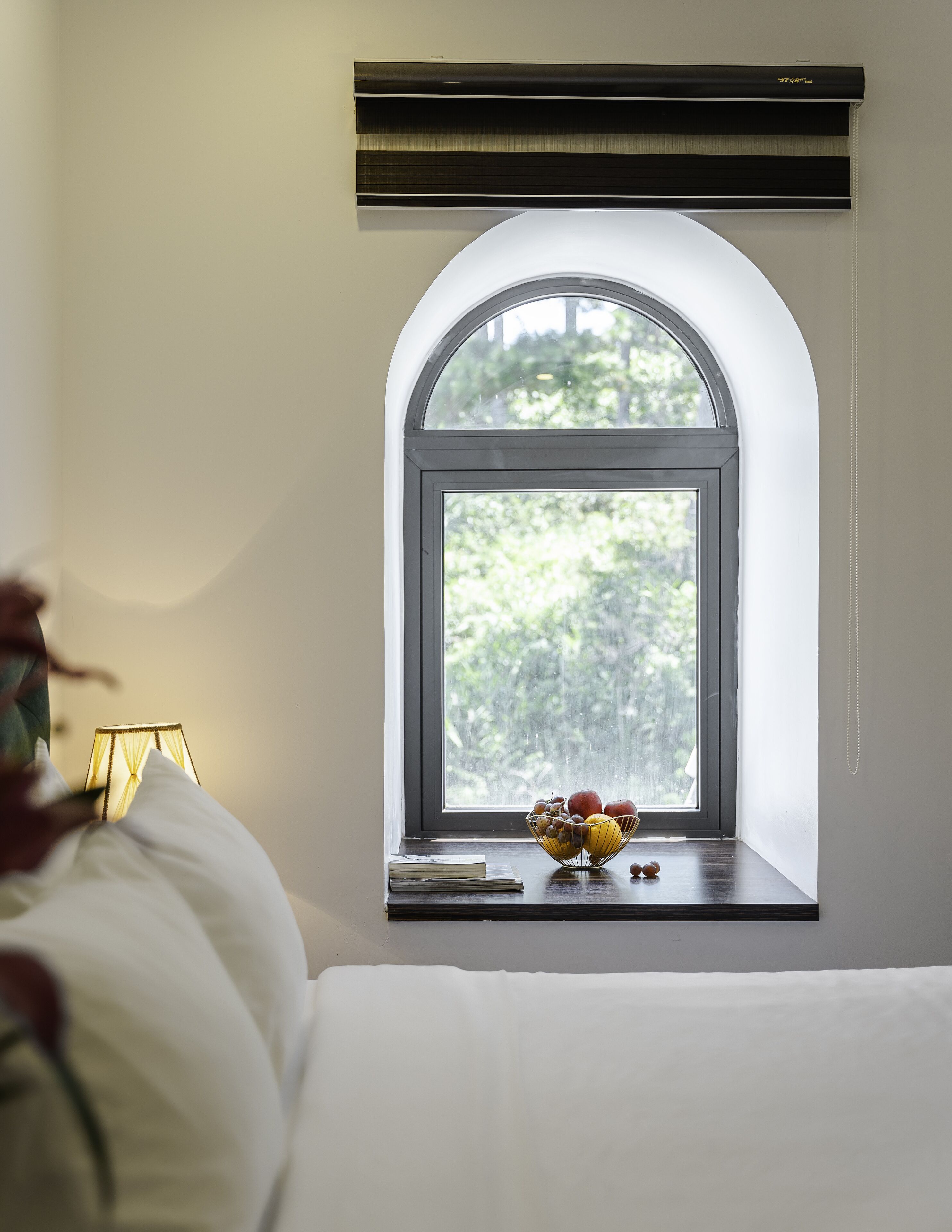 A bedroom with a large arched window, a bedside table with a bowl of fruit, and a lamp.