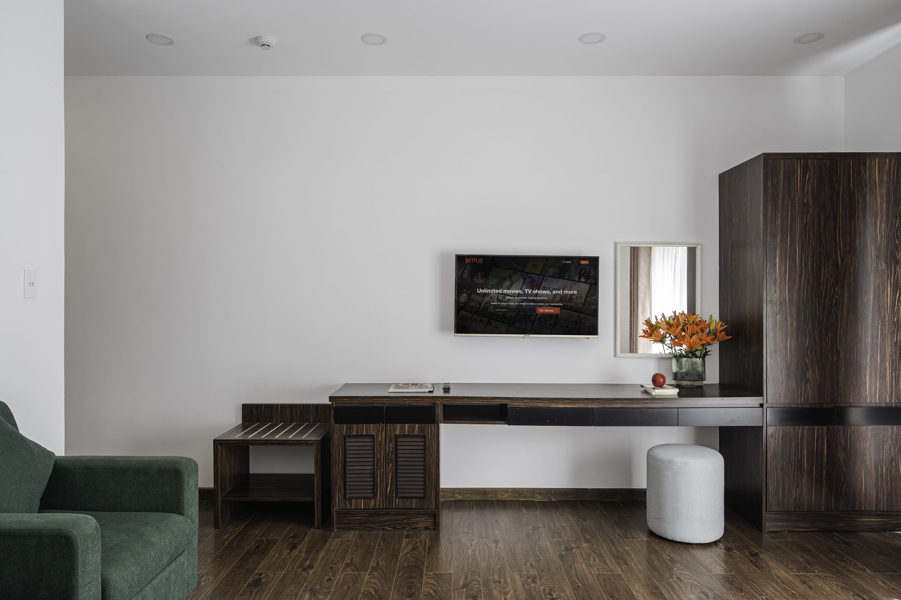 A modern living room with a wooden floor, a green armchair, a dark wooden cabinet, and a flat-screen TV mounted on the wall.
