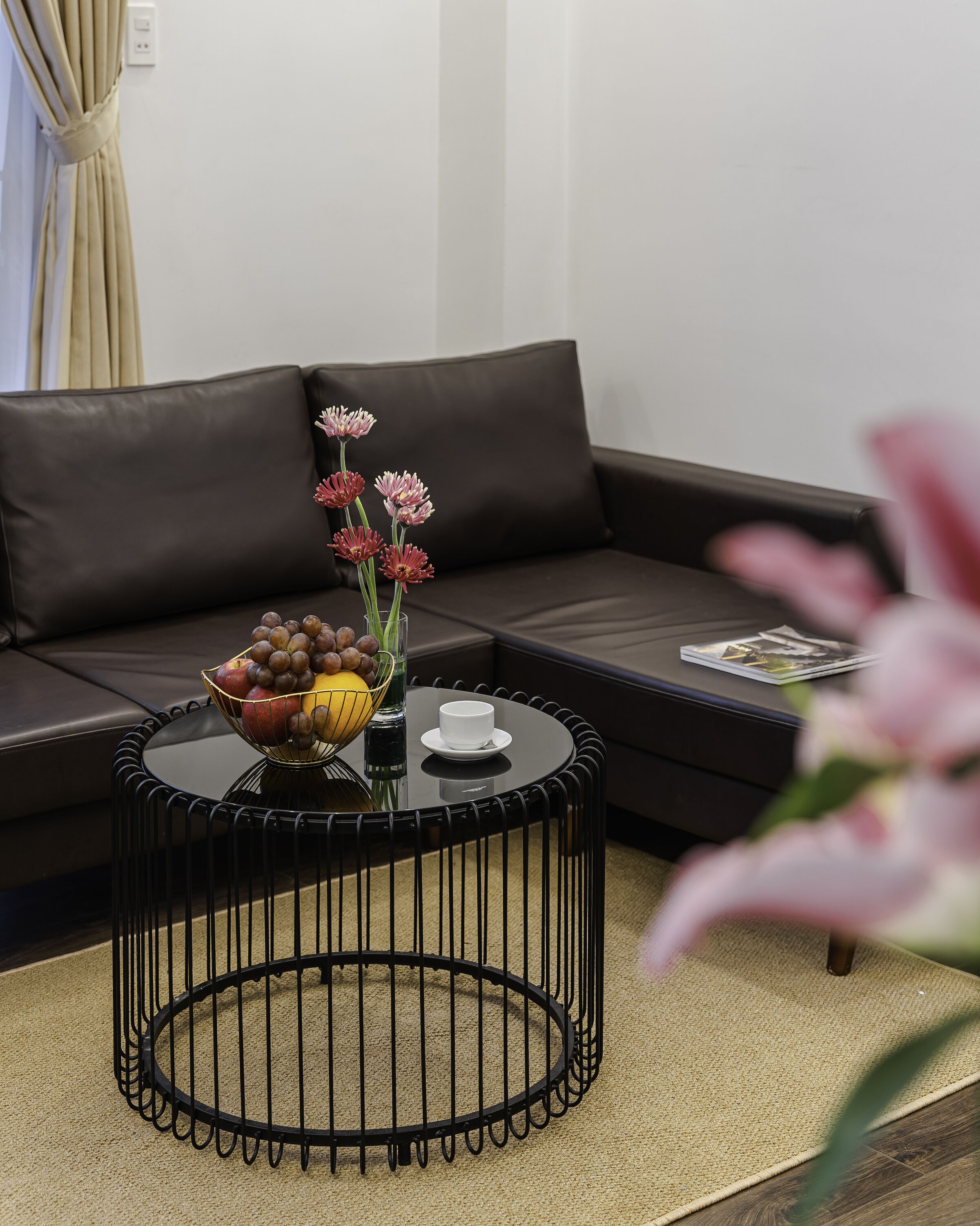 A modern living room with a black leather sofa, a round black coffee table with a fruit bowl and flowers, and a magazine on the sofa.