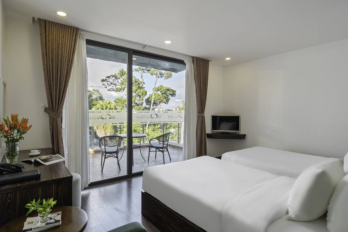 Habitación de hotel con una cama grande, un escritorio con teléfono y una revista, un televisor sobre un soporte, y un balcón con sillas y una mesa.