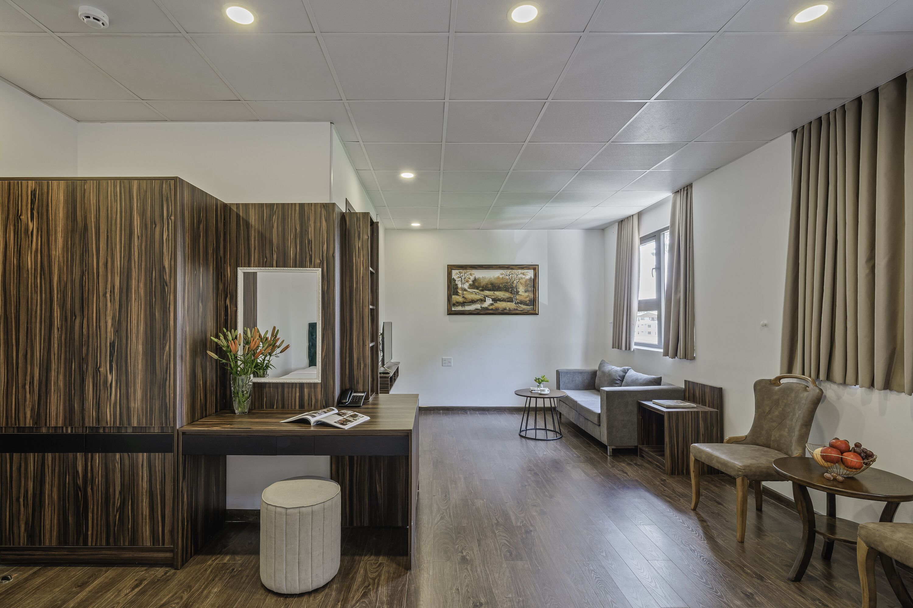 A modern hotel room with a wooden accent wall, a desk with a mirror, a gray sofa, and a dining area with a table and chairs.