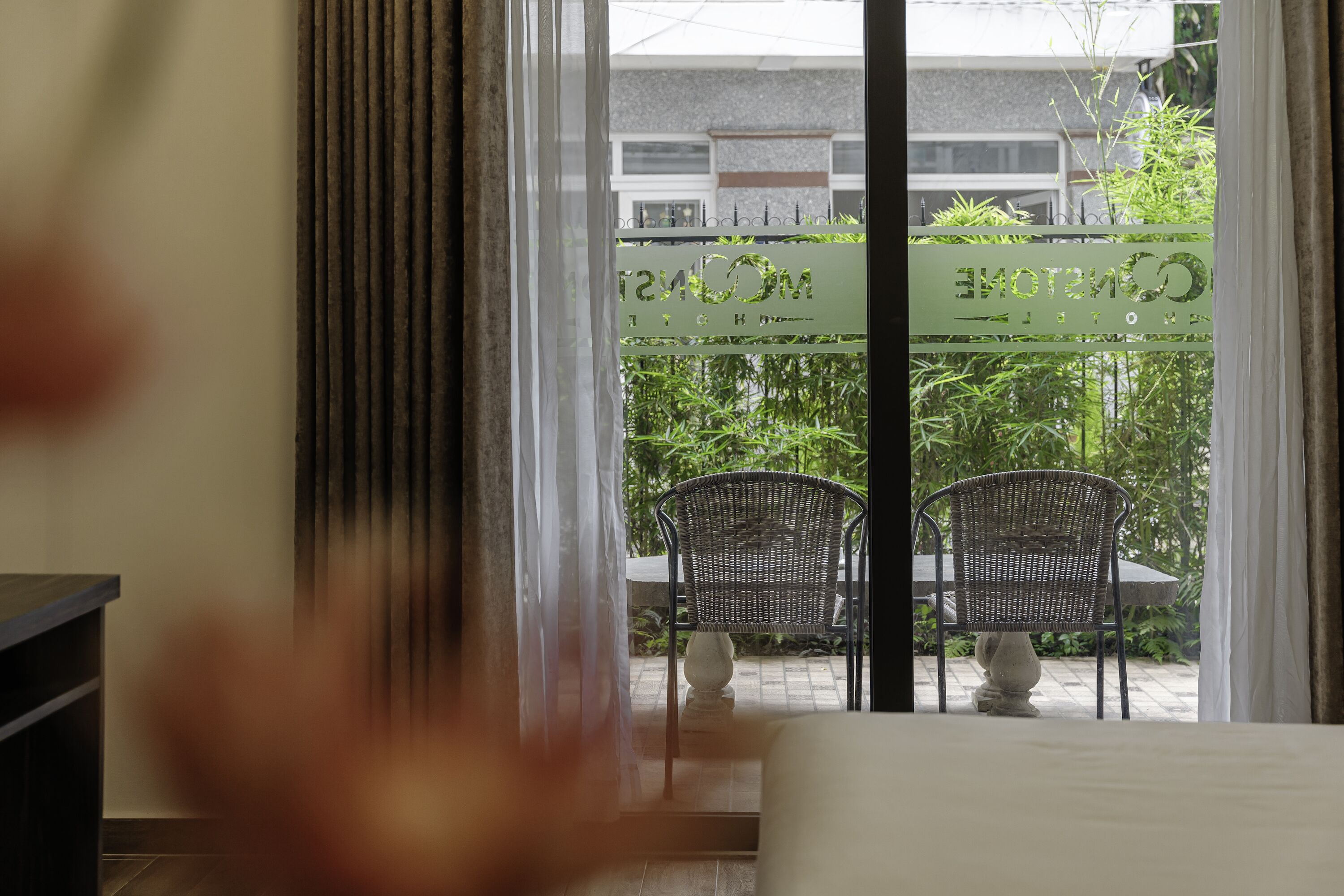 Et hotelværelse med udsigt til en patio med to fletstole og et bord, en skydedør af glas og et skilt med delvist synlig tekst.