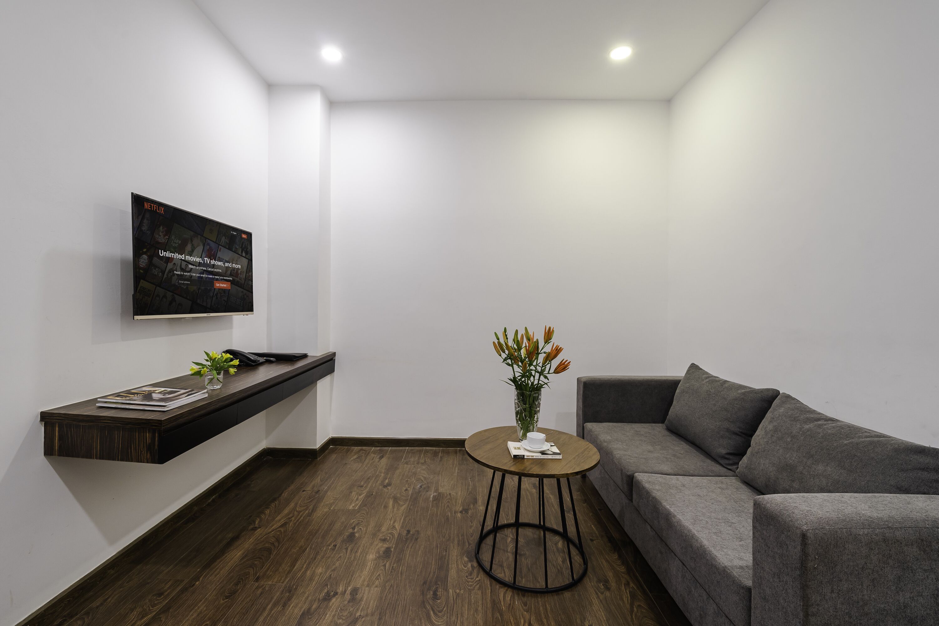 A modern living room with a grey sofa, a small round table with a vase of flowers, a mounted TV on the wall, and a wooden shelf with books and a plant.
