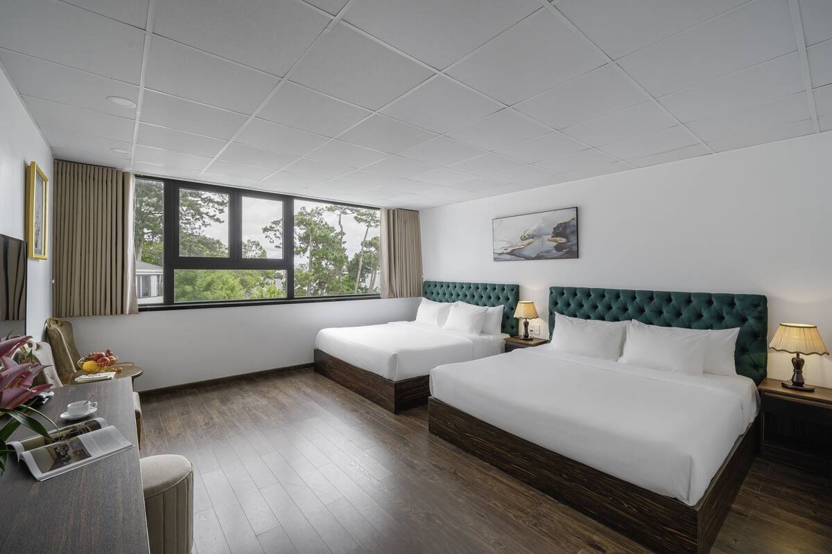 Habitación de hotel con dos camas, un escritorio, una silla, un ventanal con vistas a árboles y un cuadro en la pared.