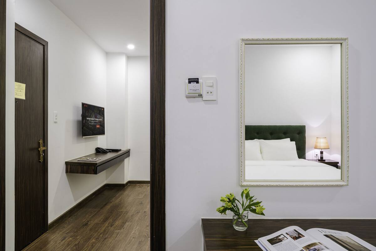 Una habitación de hotel con piso de madera oscura, una cama con sábanas blancas, una mesita de noche con lámpara, un espejo y un pequeño florero con flores.