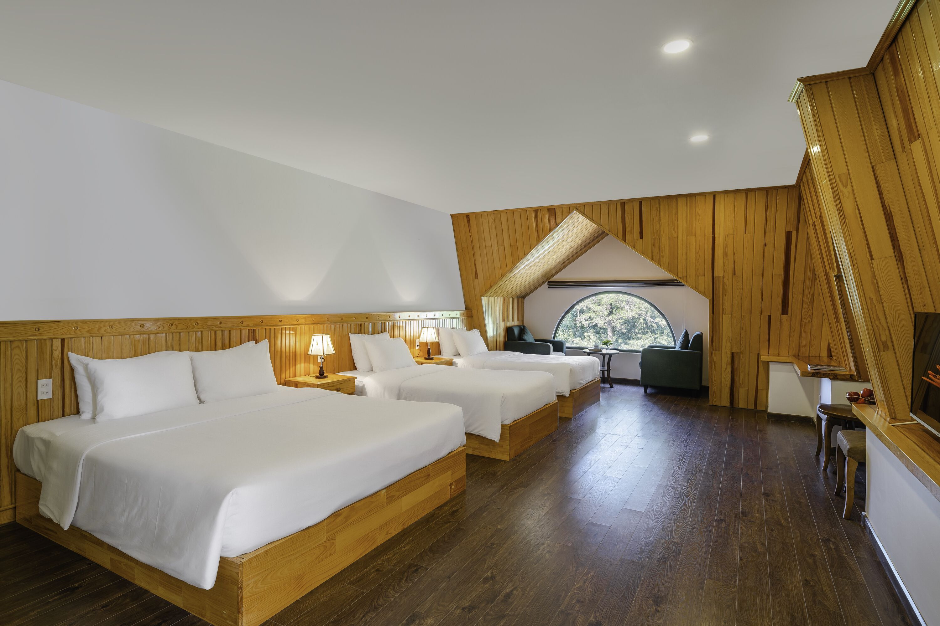 A hotel room with three wooden beds, a desk, and a view of the outdoors through a window.