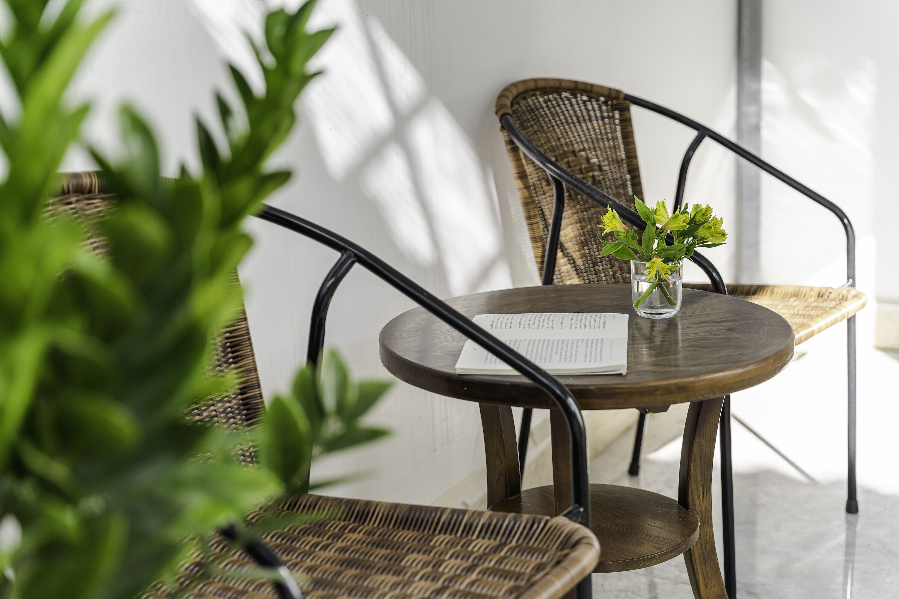 Een ronde houten tafel met een menukaart, twee rieten stoelen en een kleine vaas met groene bladeren.