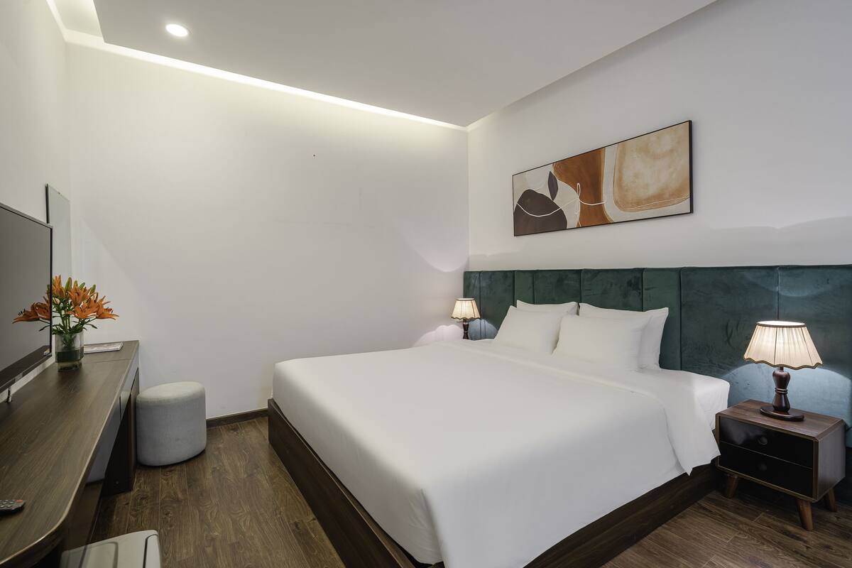 Una habitación de hotel moderna con una cama grande, una mesita de noche con lámpara, un pequeño taburete redondo, un televisor de pantalla plana y un cuadro en la pared.