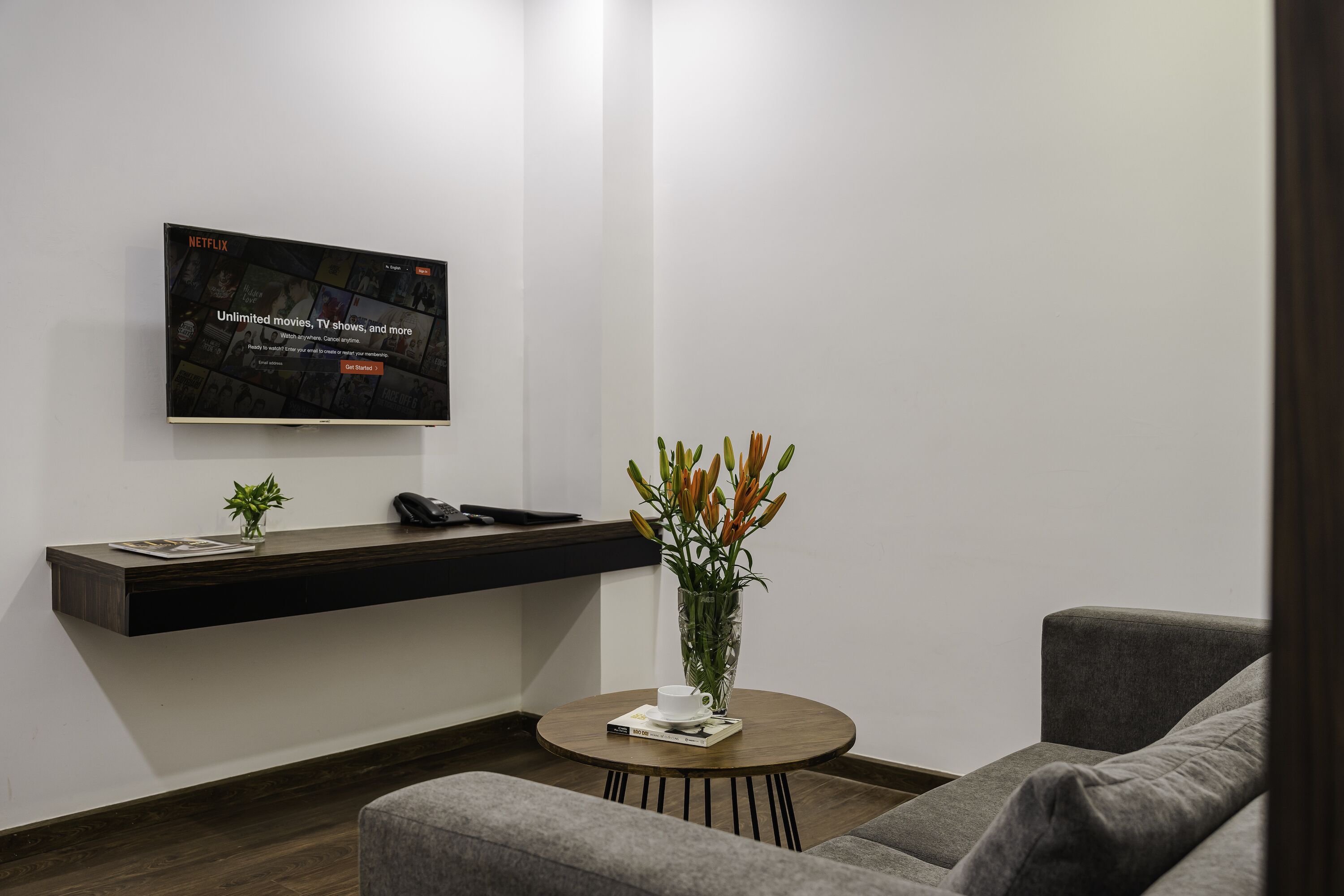 A modern living room with a mounted TV, a dark wooden shelf, a round coffee table with a book and a cup, a gray sofa, and a vase with flowers.