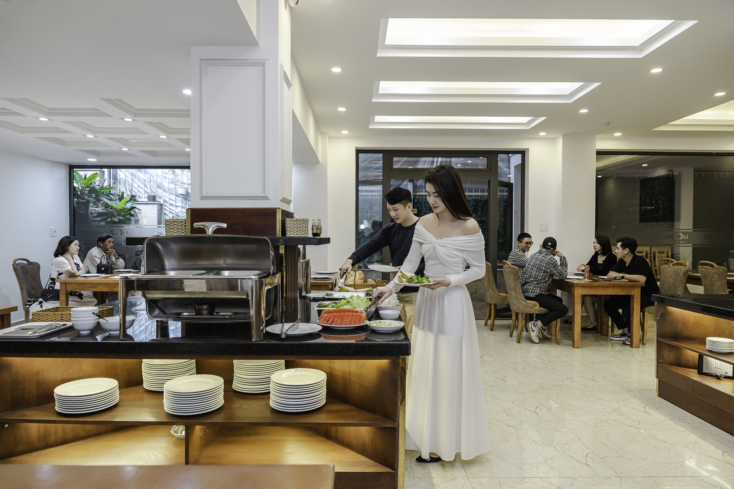 A modern dining area with a buffet setup, including a counter with plates and food, and guests seated at tables.