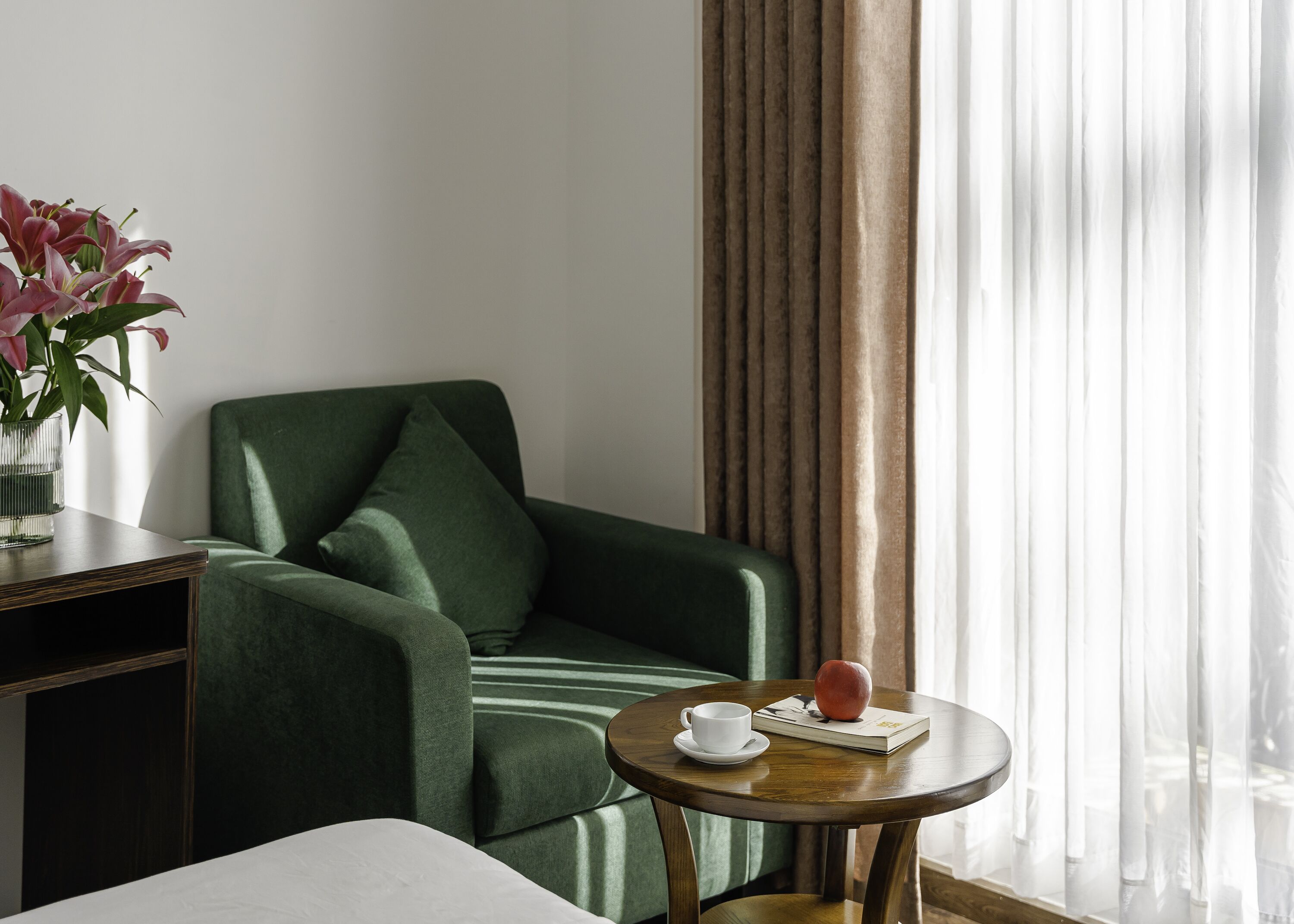 A green armchair with a cushion, a small round wooden table with a white cup and a book, and a vase with flowers.