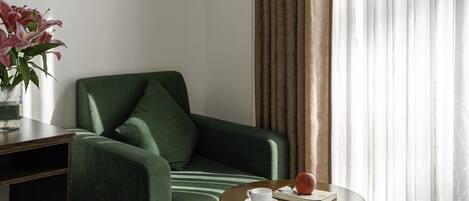 A green armchair with a cushion, a small round wooden table with a white cup and a book, and a vase with flowers.