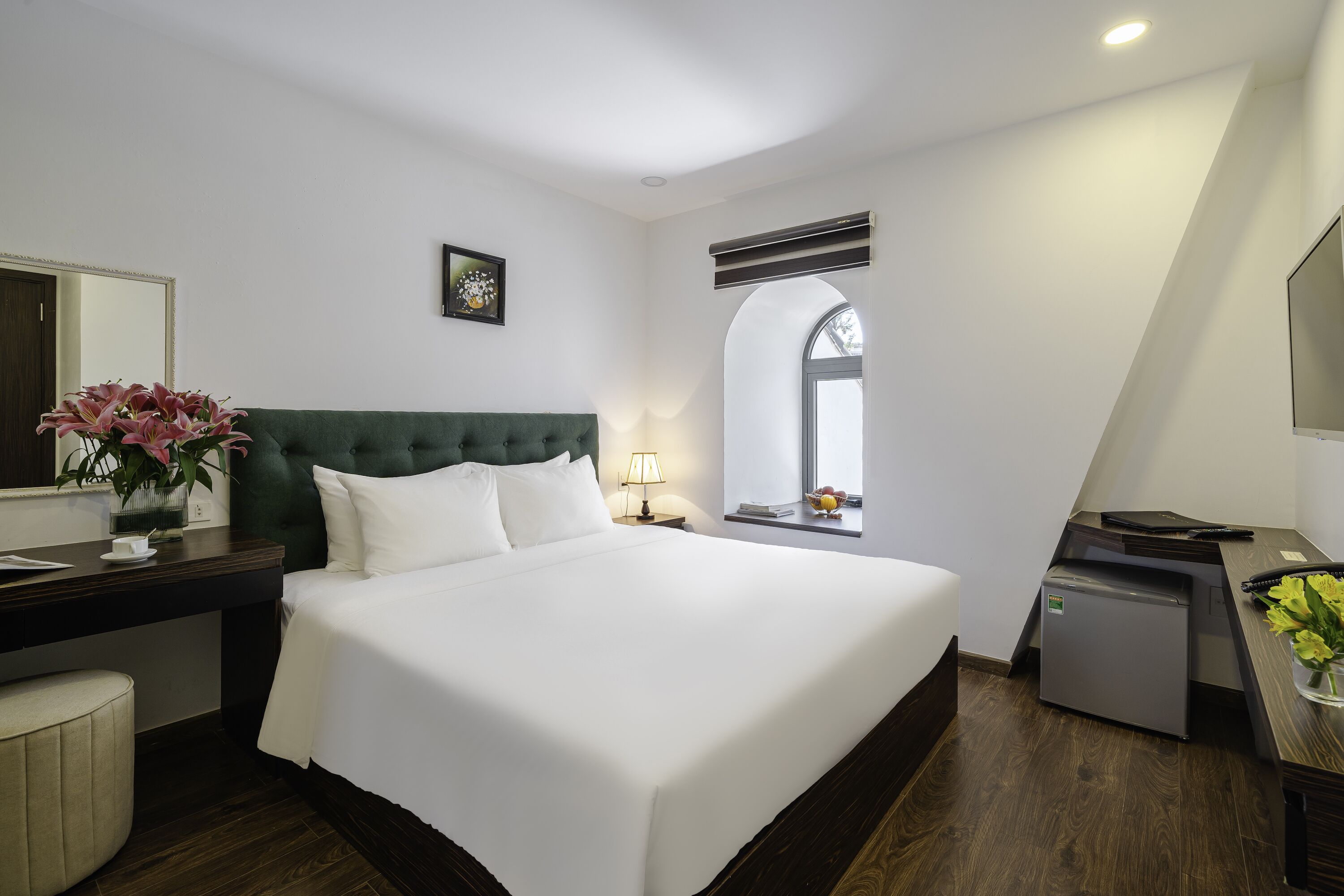 A hotel room with a large bed, a desk with a computer, a small fridge, a window with a view, and a mirror.