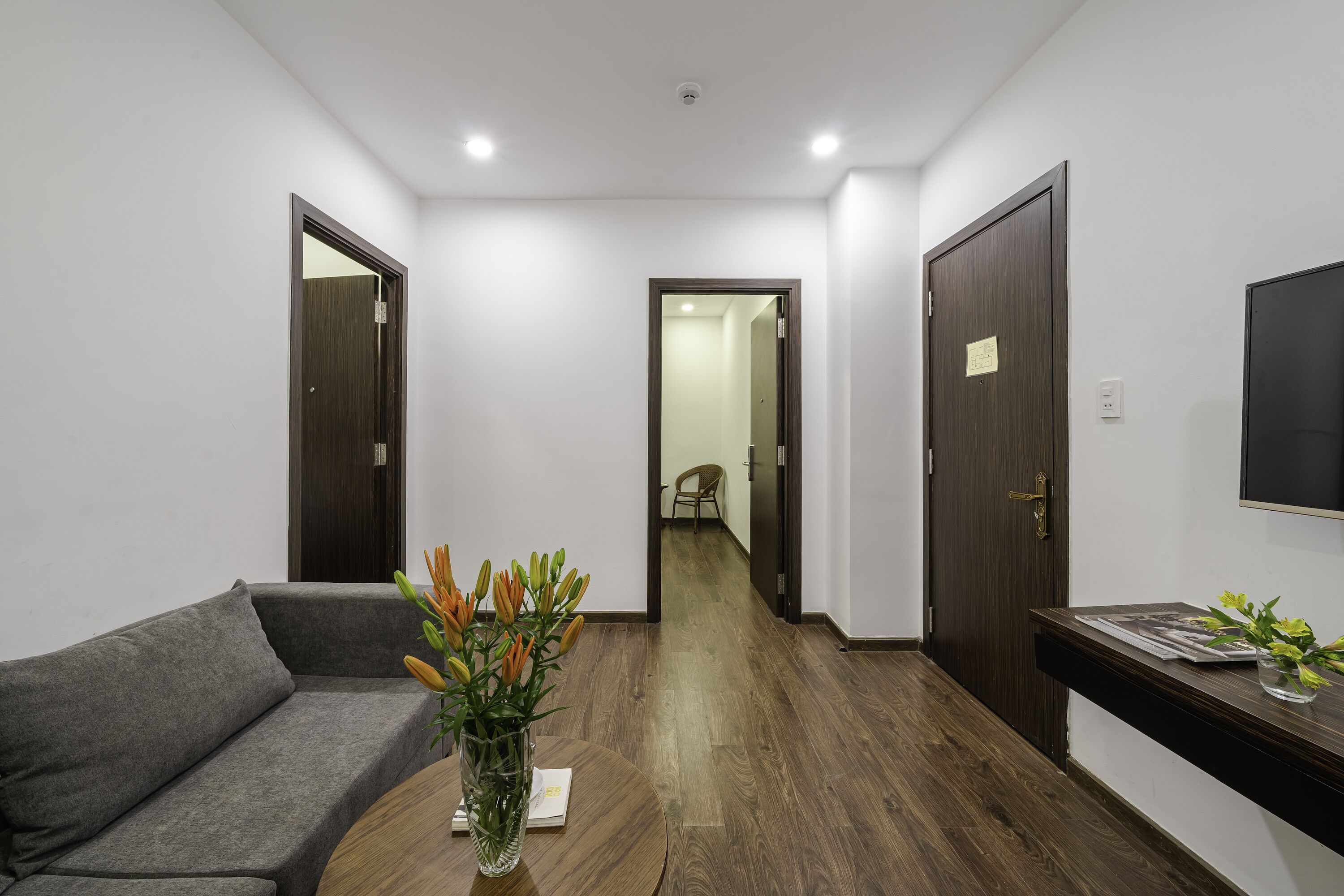 Un interior moderno con un sofá gris, una mesa de centro de madera y un jarrón con flores, y un televisor montado en la pared.