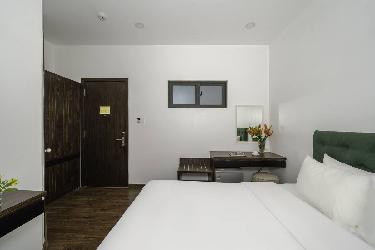 Habitación de hotel con cama, escritorio, silla, refrigerador pequeño, espejo y puerta.