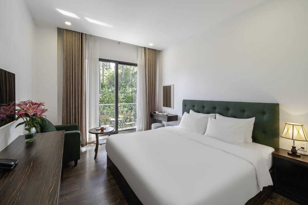 Habitación de hotel con una cama grande, un escritorio con una silla, una mesita de noche con lámpara y vistas a zonas verdes por la ventana.