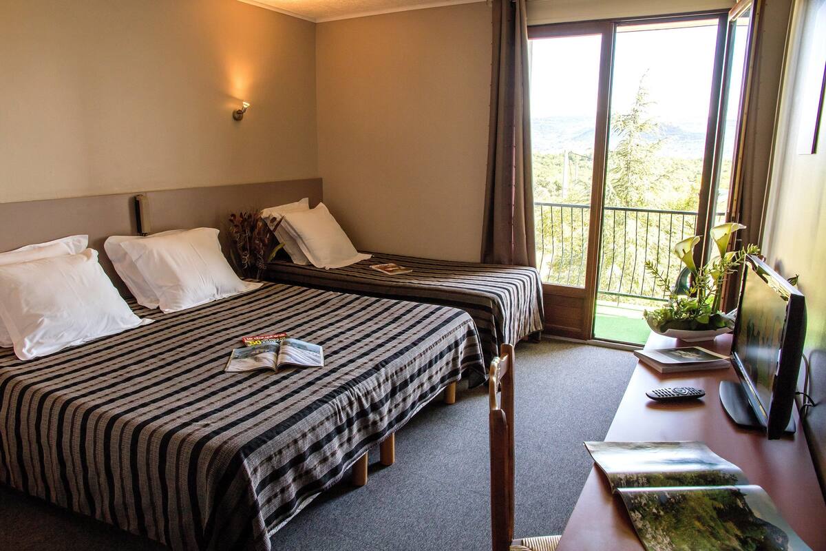 Habitación de hotel con dos camas, un escritorio con computadora y televisión, y un balcón con vistas.