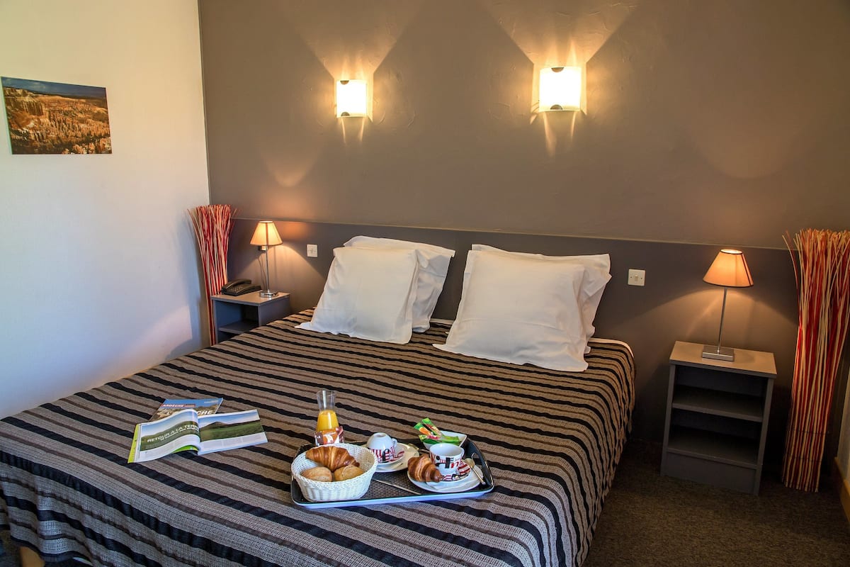 Habitación de hotel con una cama, dos mesitas de noche, una lámpara y una bandeja de desayuno con croissants, una botella y un libro.