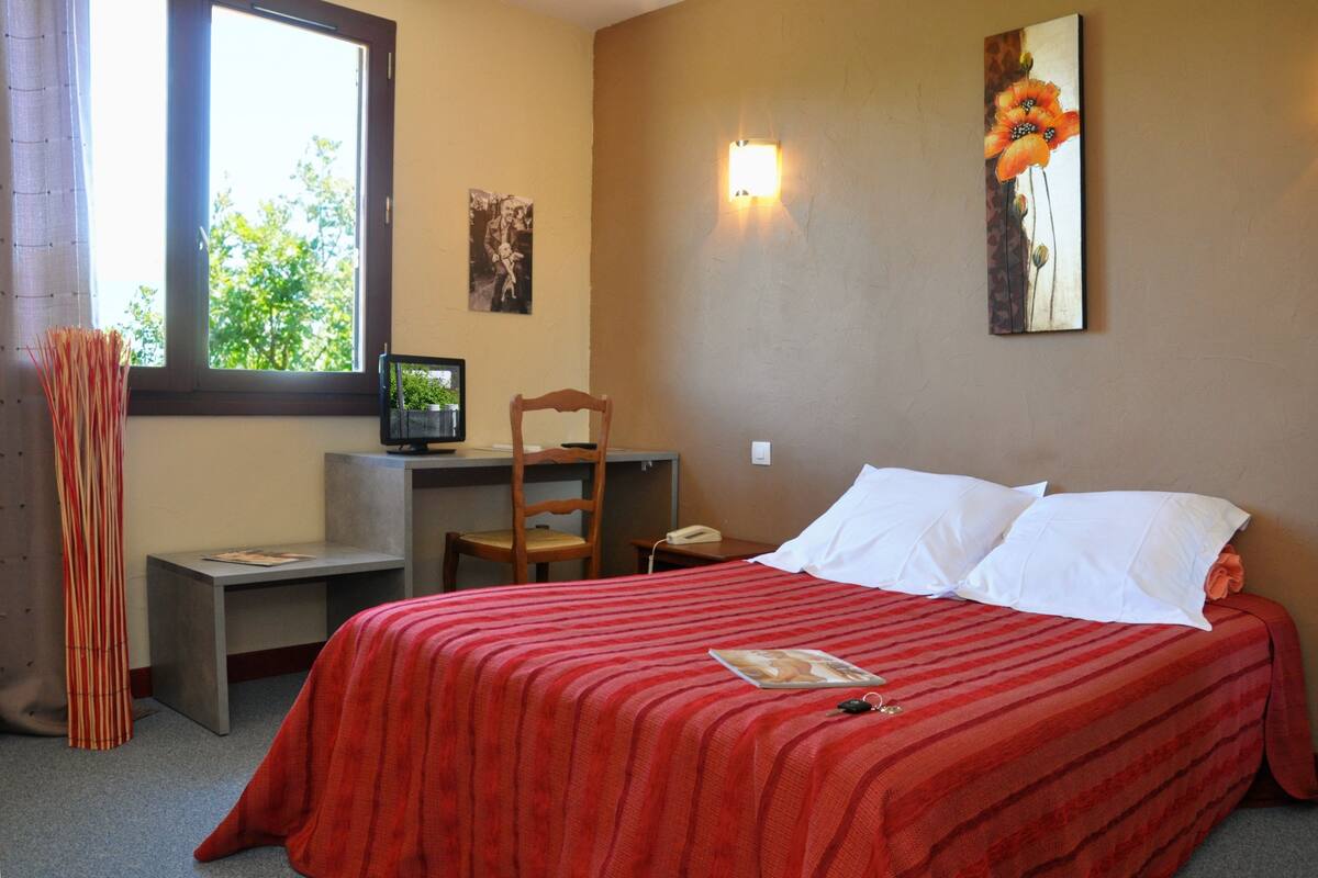 Un dormitorio con cama, escritorio, silla, ventana y una lámpara fijada a la pared.