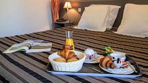 A neatly made bed with white pillows, a bedside lamp, a tray with croissants, a bottle of orange juice, a cup of coffee, and a book on a striped bedspread. - Hotel Les Persedes Sud Ardeche (Lavilledieu)