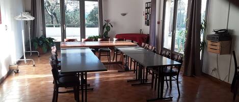 A conference room with a long table, chairs, and a projector. There are windows with curtains, a plant, and a fire extinguisher on the wall.