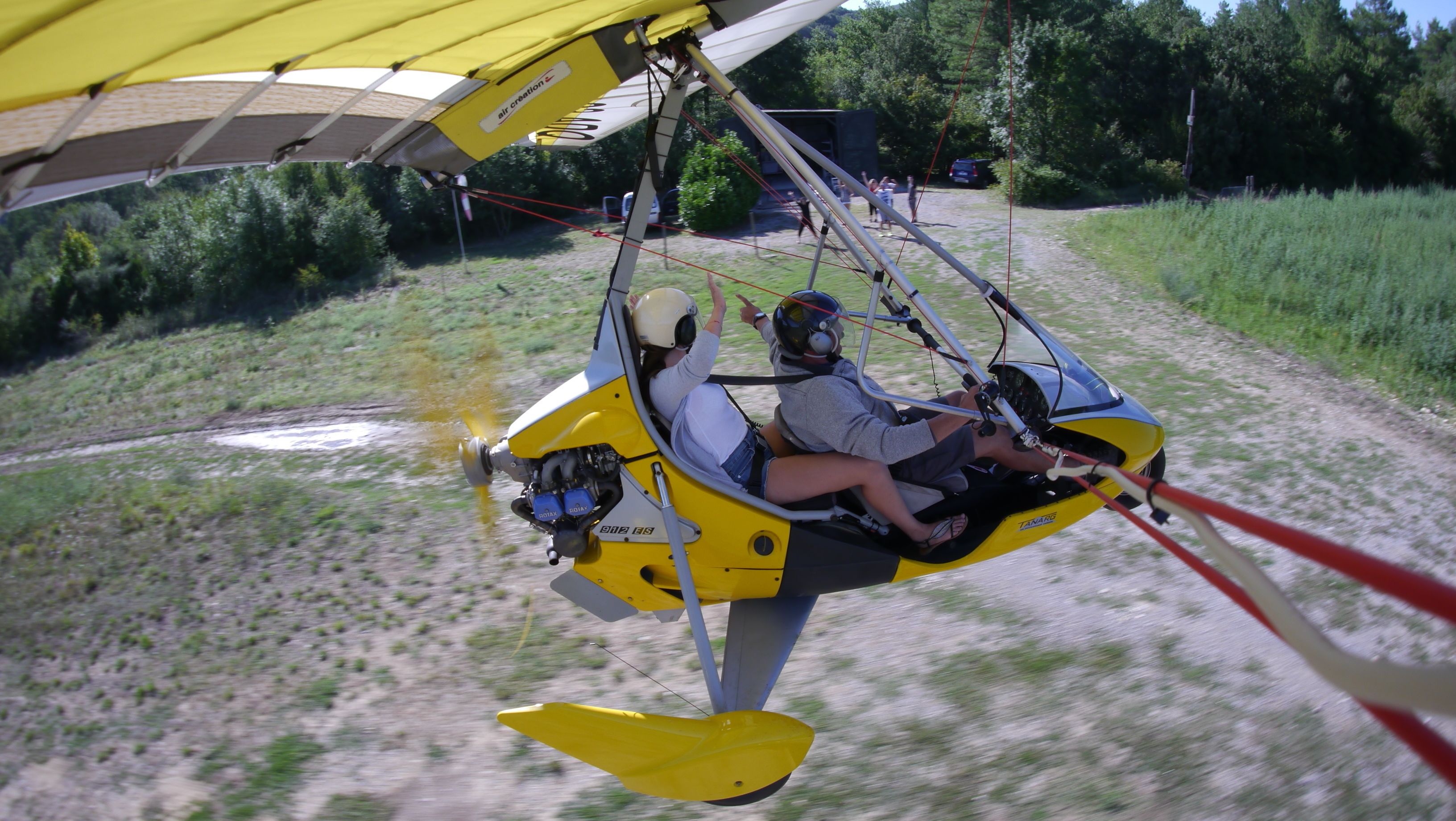 A yellow hang glider with two passengers, one wearing a helmet, flying over a dirt path with greenery on either side.