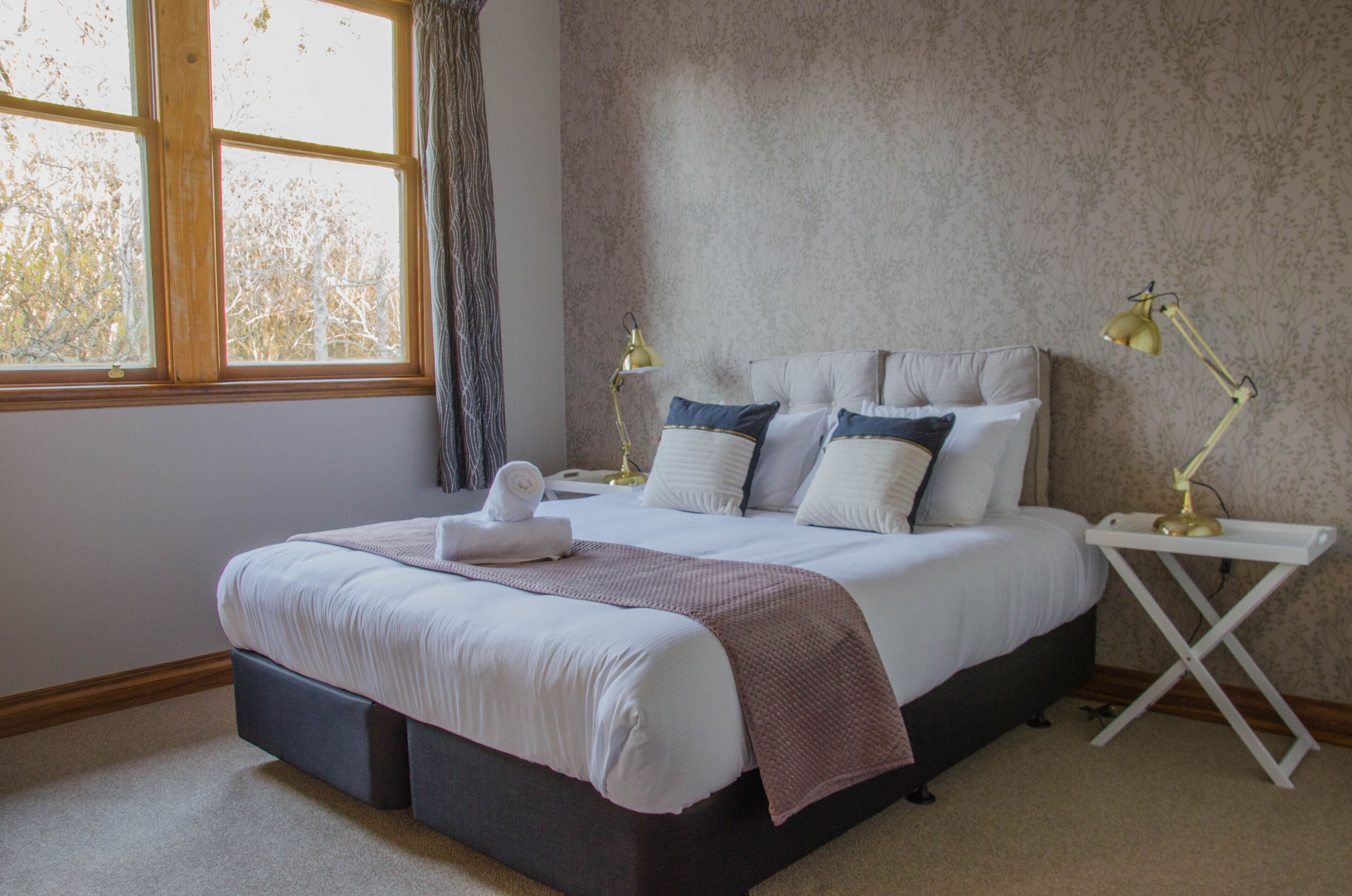 A neatly made bed with pillows and a blanket, a bedside table with a lamp, and a window with curtains.