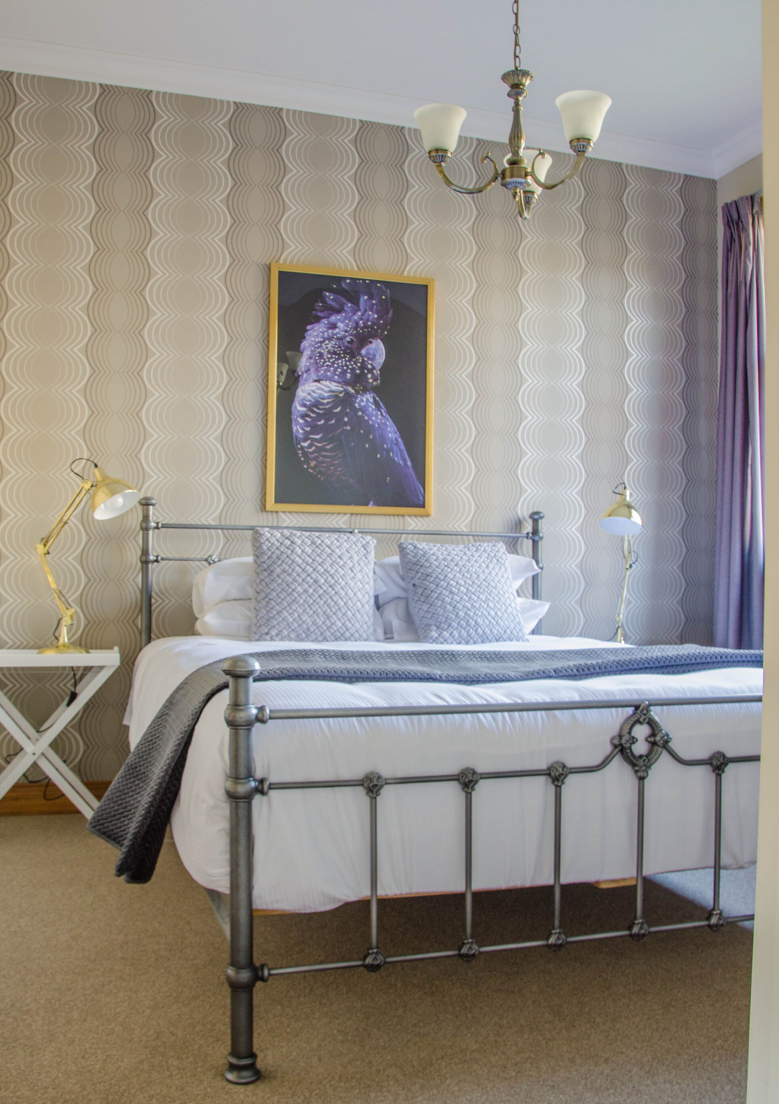 A neatly arranged bedroom with a metal bed frame, a bedside table with a lamp, a wall-mounted painting, and a patterned wallpaper.
