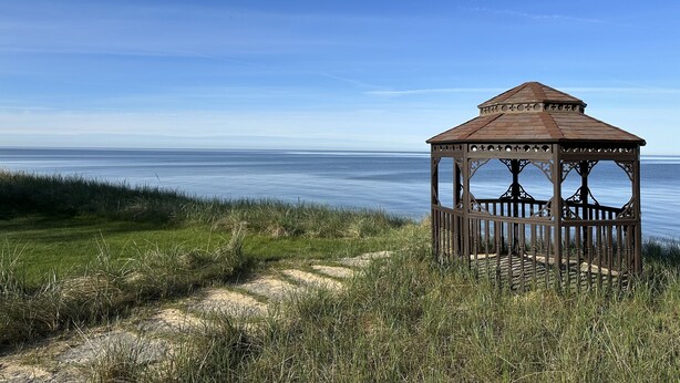 A wooden gazebo with intricate designs, situated on a grassy path by a calm body of water under a clear blue sky. - Paradise Lake Michigan Beach home with inground pool,  hot tub, sleeps 20 (Norton Shores)