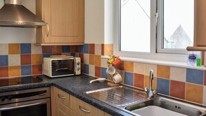 A kitchen with wooden cabinets, a microwave, a stove, a sink, and a window with a view of a building. - 1 bedroom accommodation in Bowness-on-Windermere (Bowness-on-Windermere)