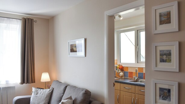 A modern living room with a grey sofa, a lamp, and a kitchen visible through an open door. - 1 bedroom accommodation in Bowness-on-Windermere (Bowness-on-Windermere)