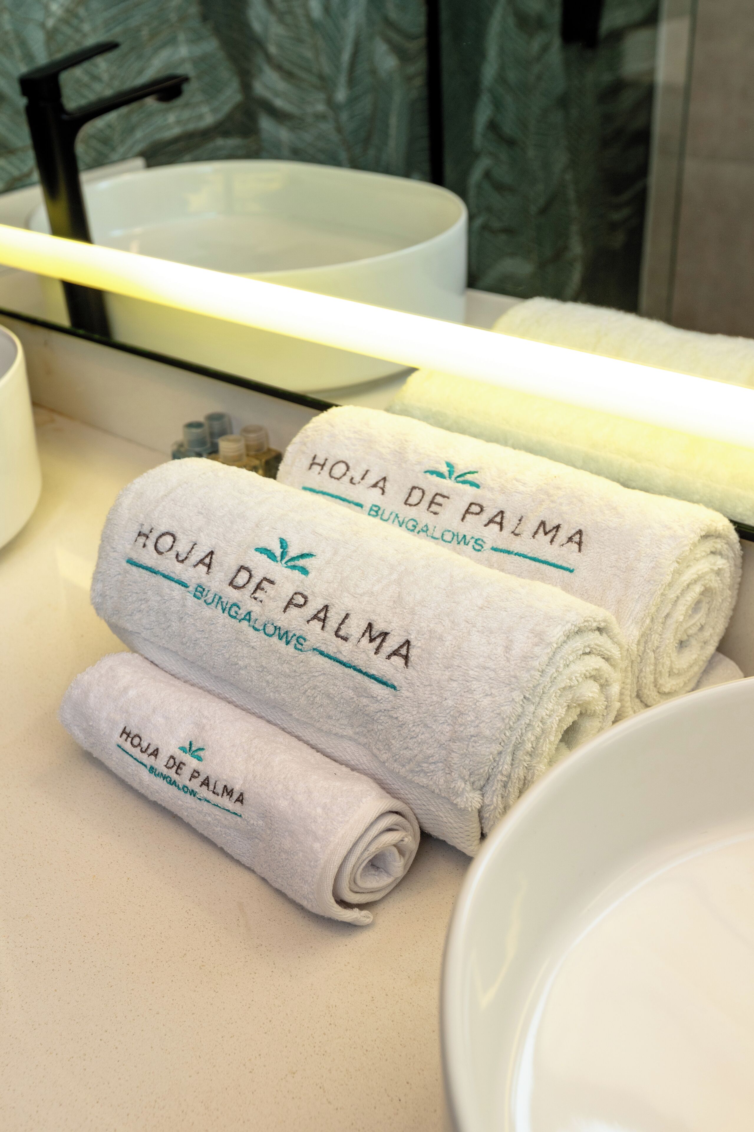 A bathroom with a white sink, a mirror, and three towels rolled up with the text "HOJA DE PALMA BUNGALOWS" on them.